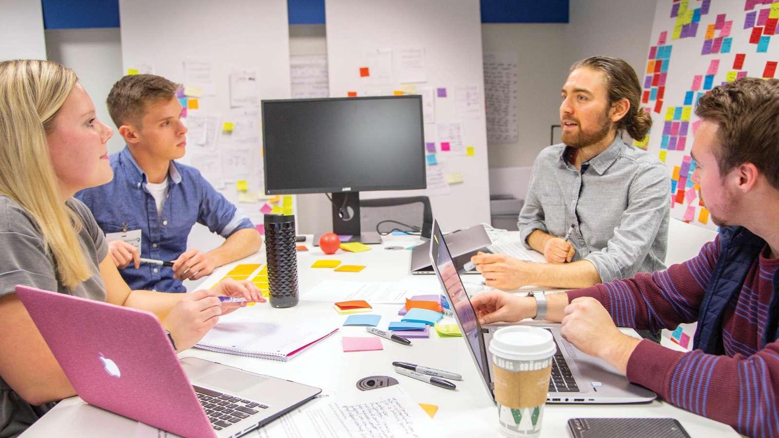 Four students meet at a table in a design lab surrounded with computers and project notes